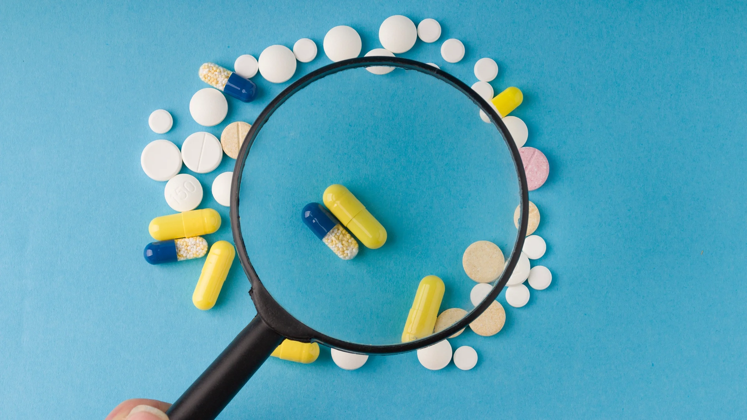 Magnifying glass held up to a pile of pills on a bright blue background.
