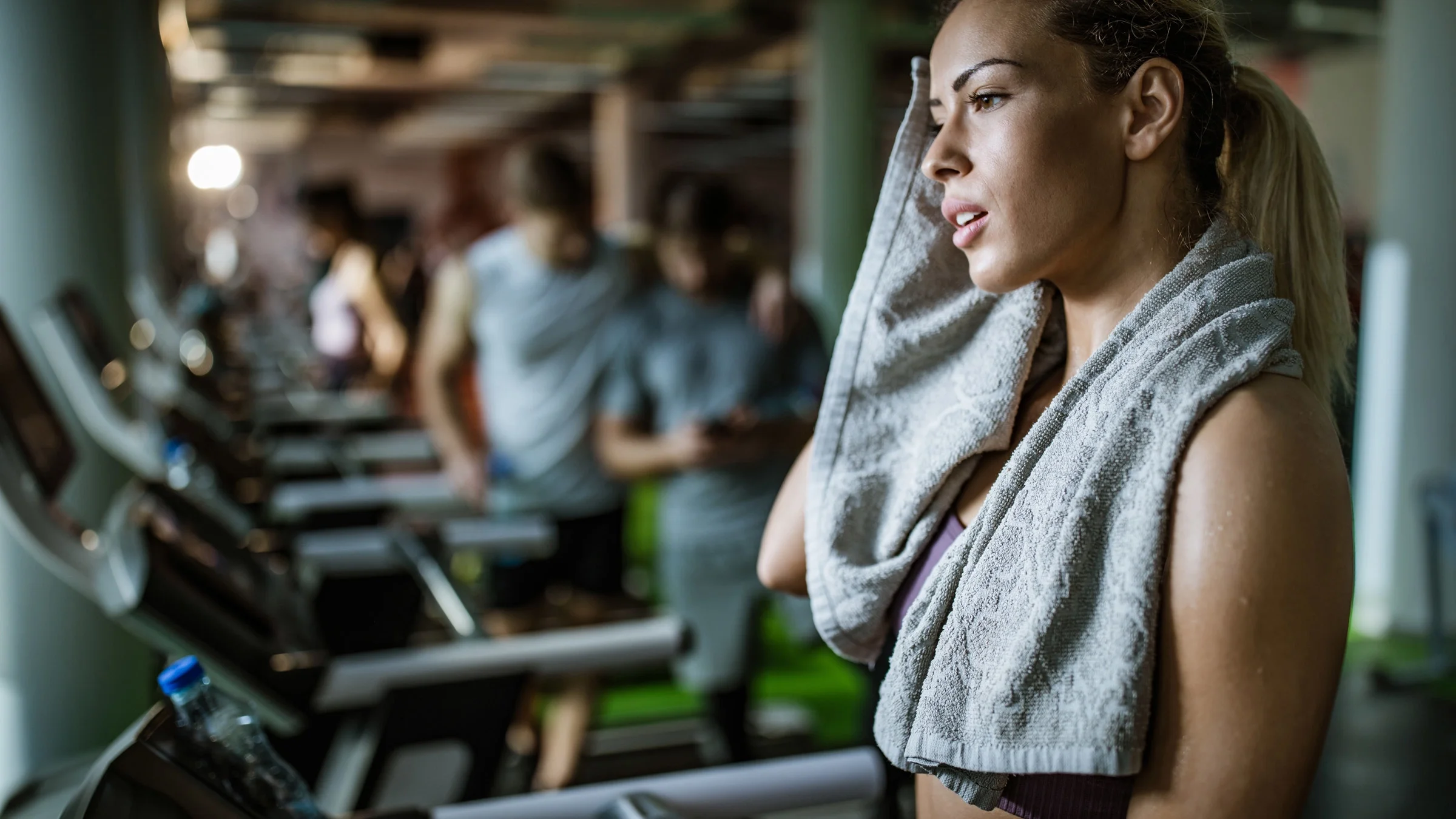 Woman with a ponytail is on a treadmill at the gym. She is using a tan, hand towel to wipe away her sweat. The background of the gym is blurred.