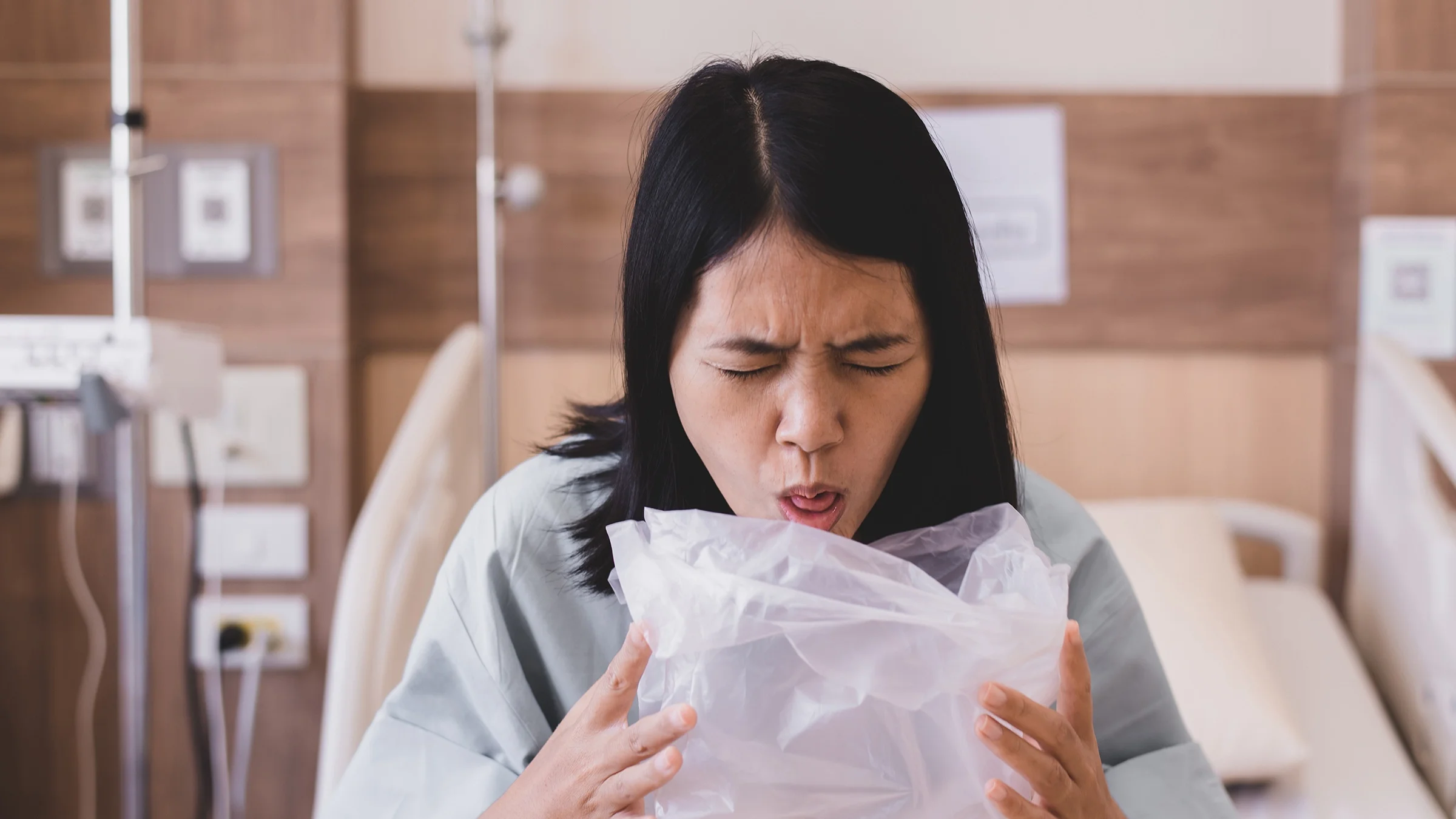 Woman sitting up in a hospital bed vomiting into a plastic bag.