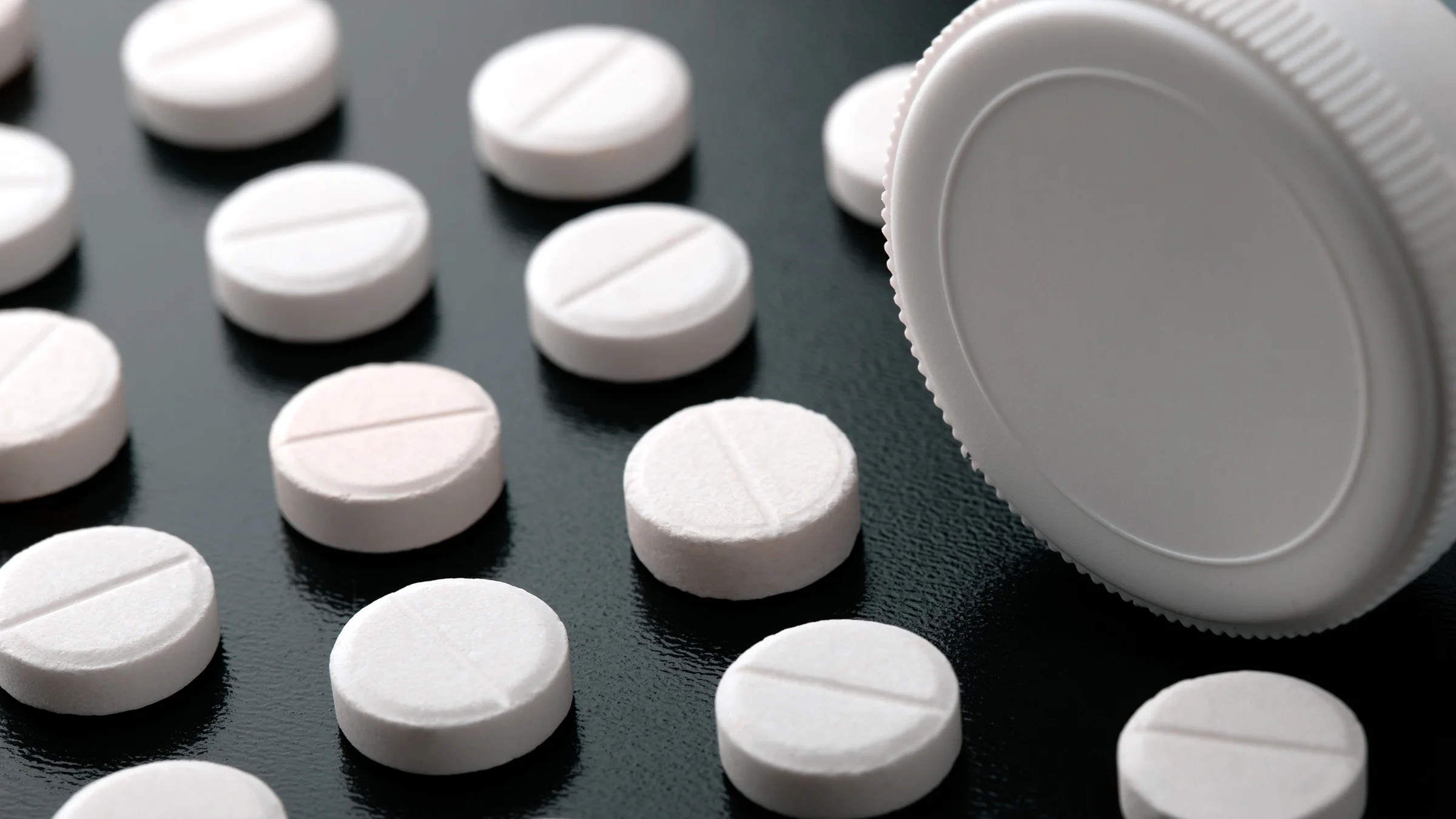 White, round pills creating a diagonal pattern on a black background. There is a white pill bottle in the upper-right corner laying on its side.