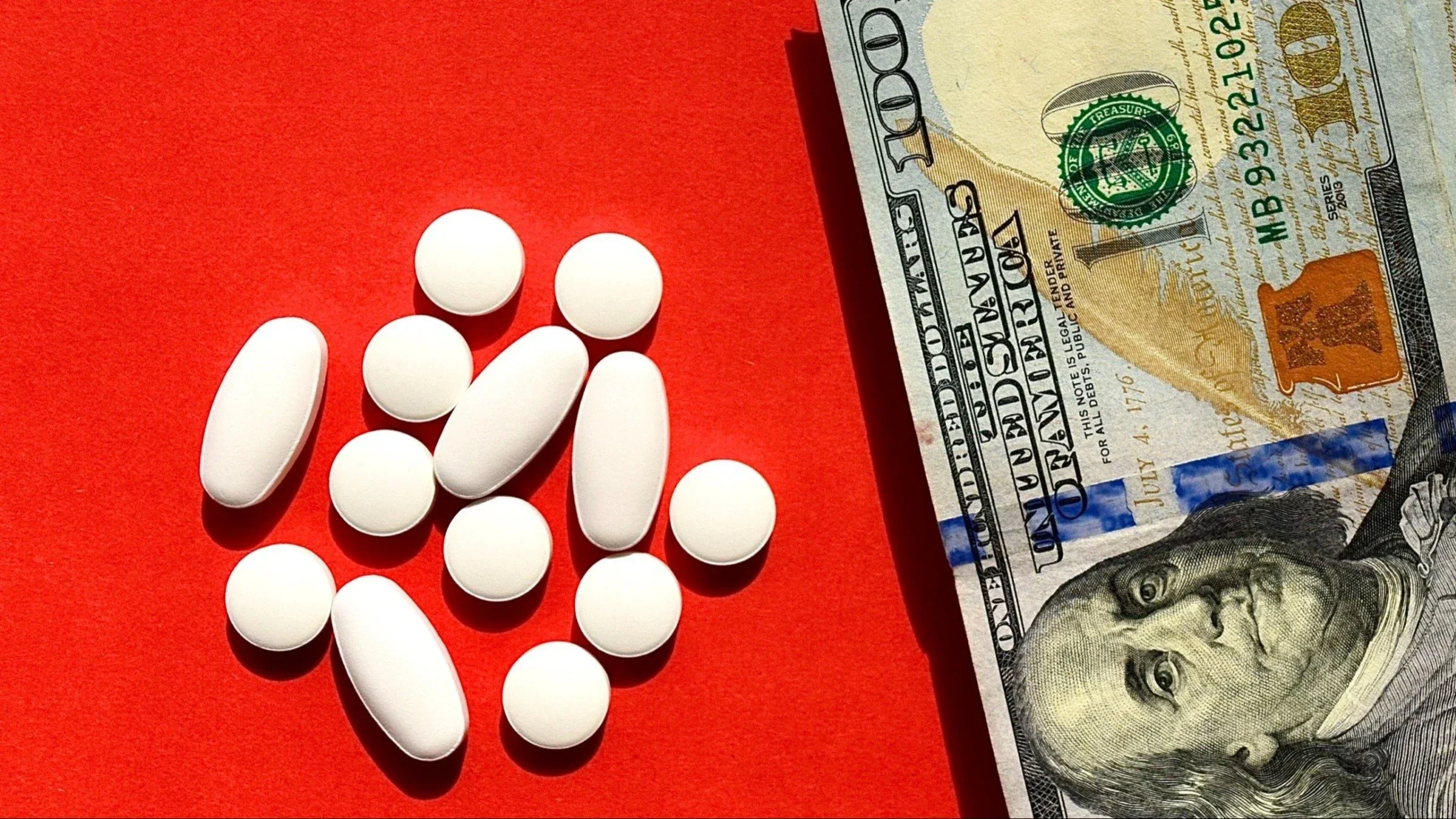 A pile of various shaped white pills next to a hundred dollar bill on a bright red background.
Viktoriya Fivko/iStock via Getty Images
