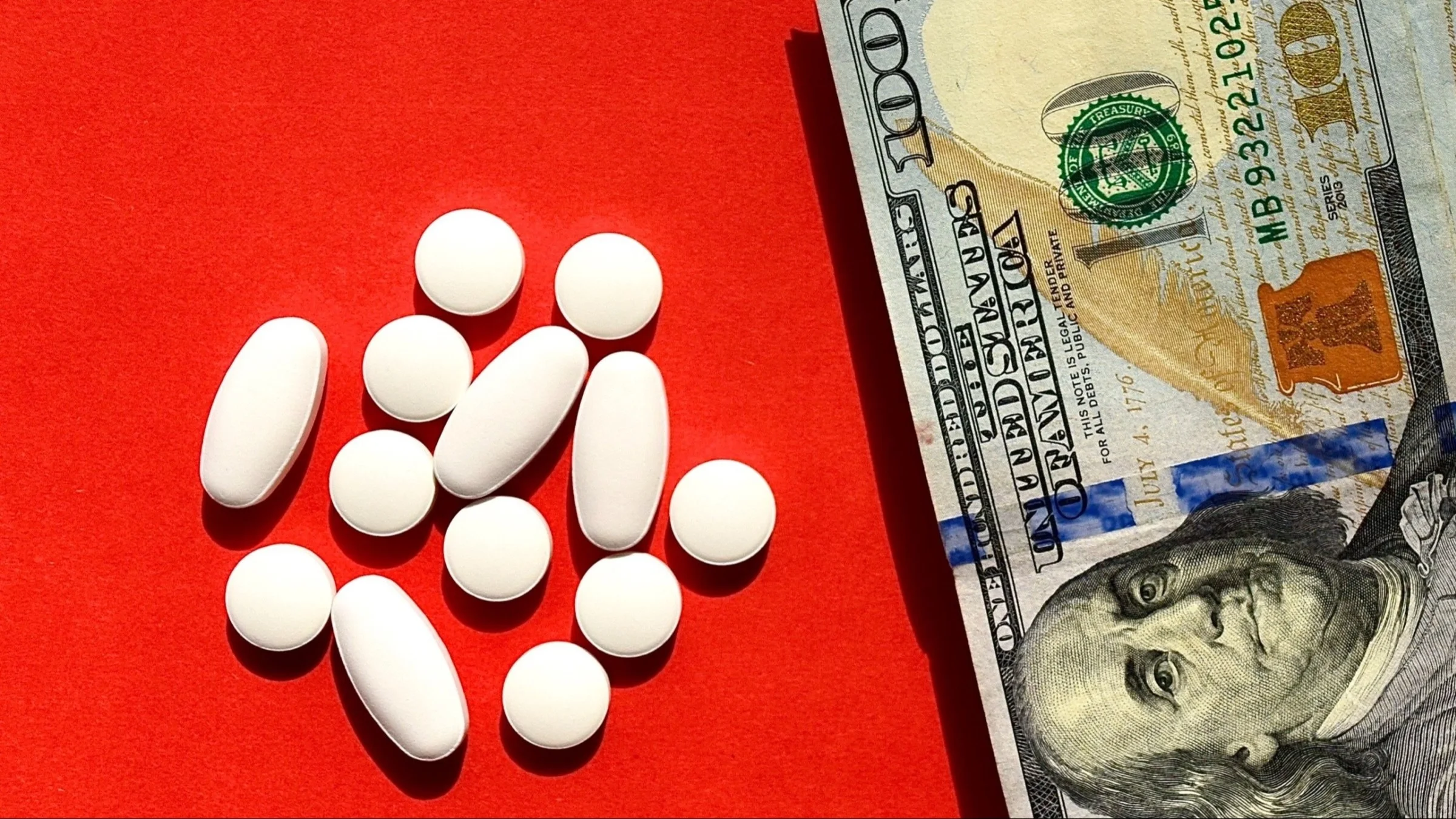 A pile of various shaped white pills next to a hundred dollar bill on a bright red background.