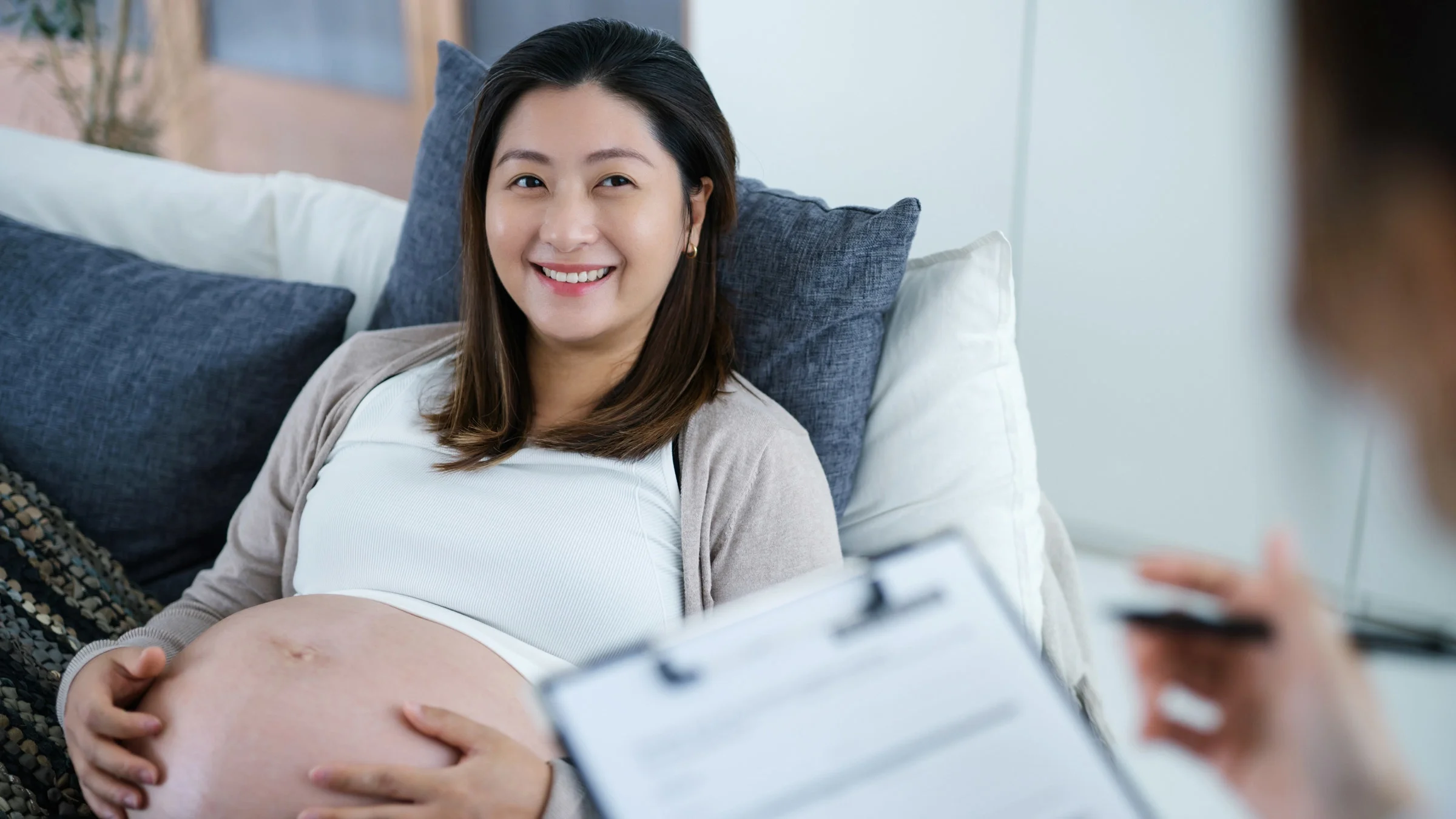 A pregnant person sitting comfortably at home, talking to a midwife who is taking notes on a clipboard.
