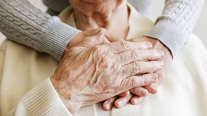 Close-up cropped shot of an older adult’s hands interlaced with a nurse's hands. They’re both resting their hands on the older adult’s chest.