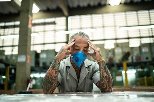 A man sitting at his work bench at an industrial job with his hands on his temples looking sad and worried.