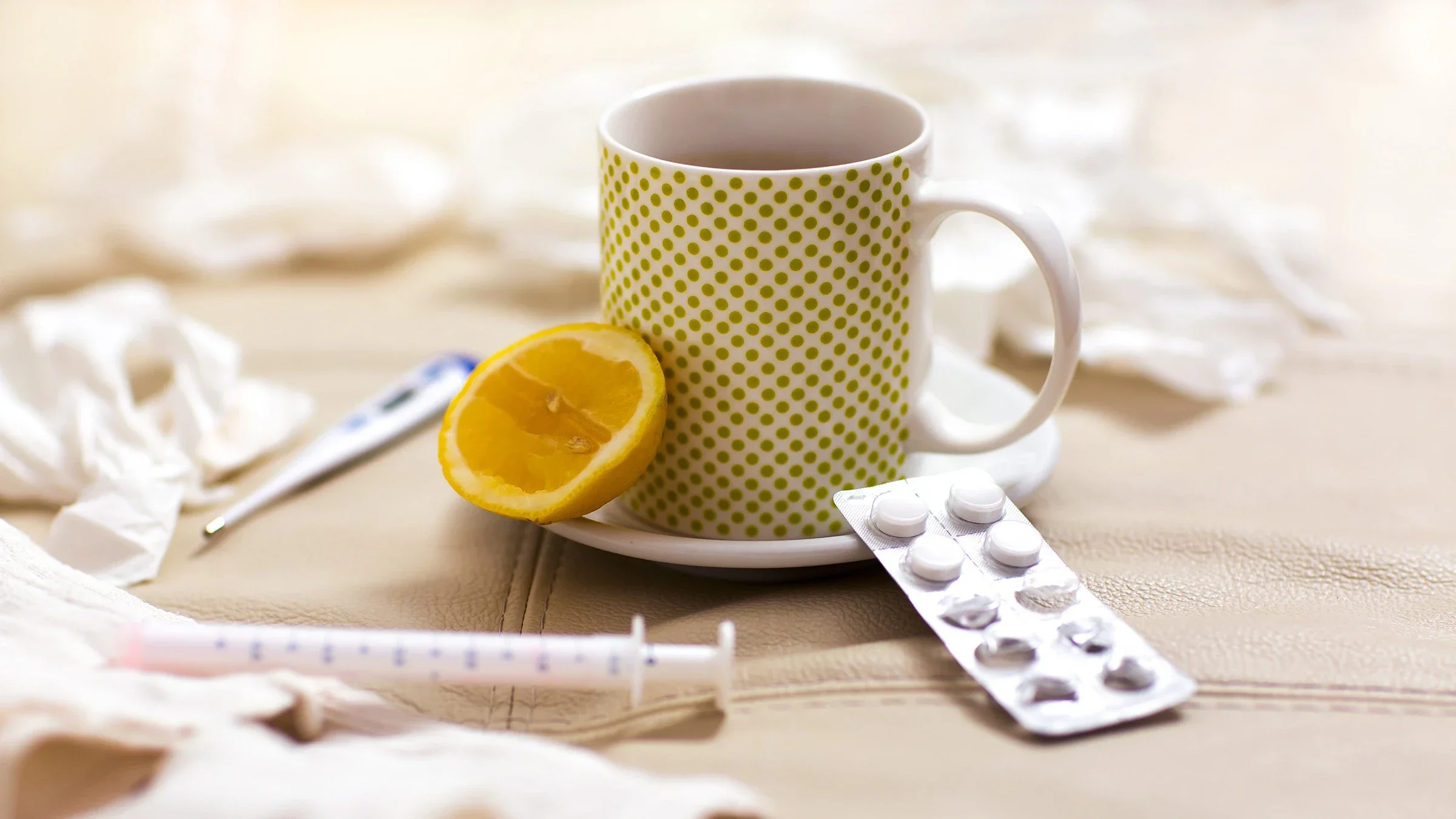 White mug and saucer with green polka dots with a lemon wedge placed on a couch. There are other cold and flu items surrounding it including: tissues, thermometer, medicine blister pack, and syringe.
Jovanmandic/iStock via Getty Images