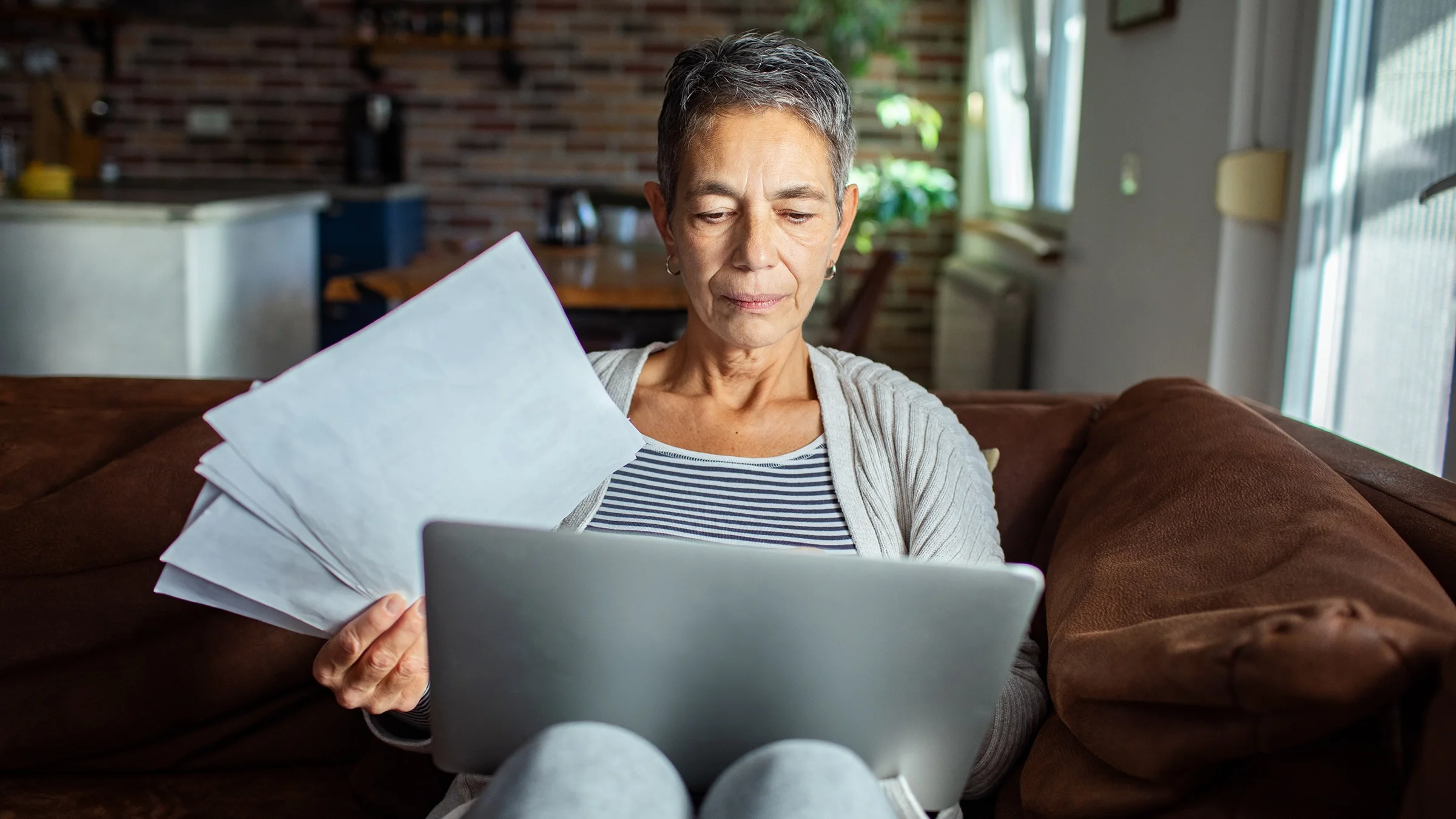 Woman using laptop on a couch at home while reviewing paperwork.