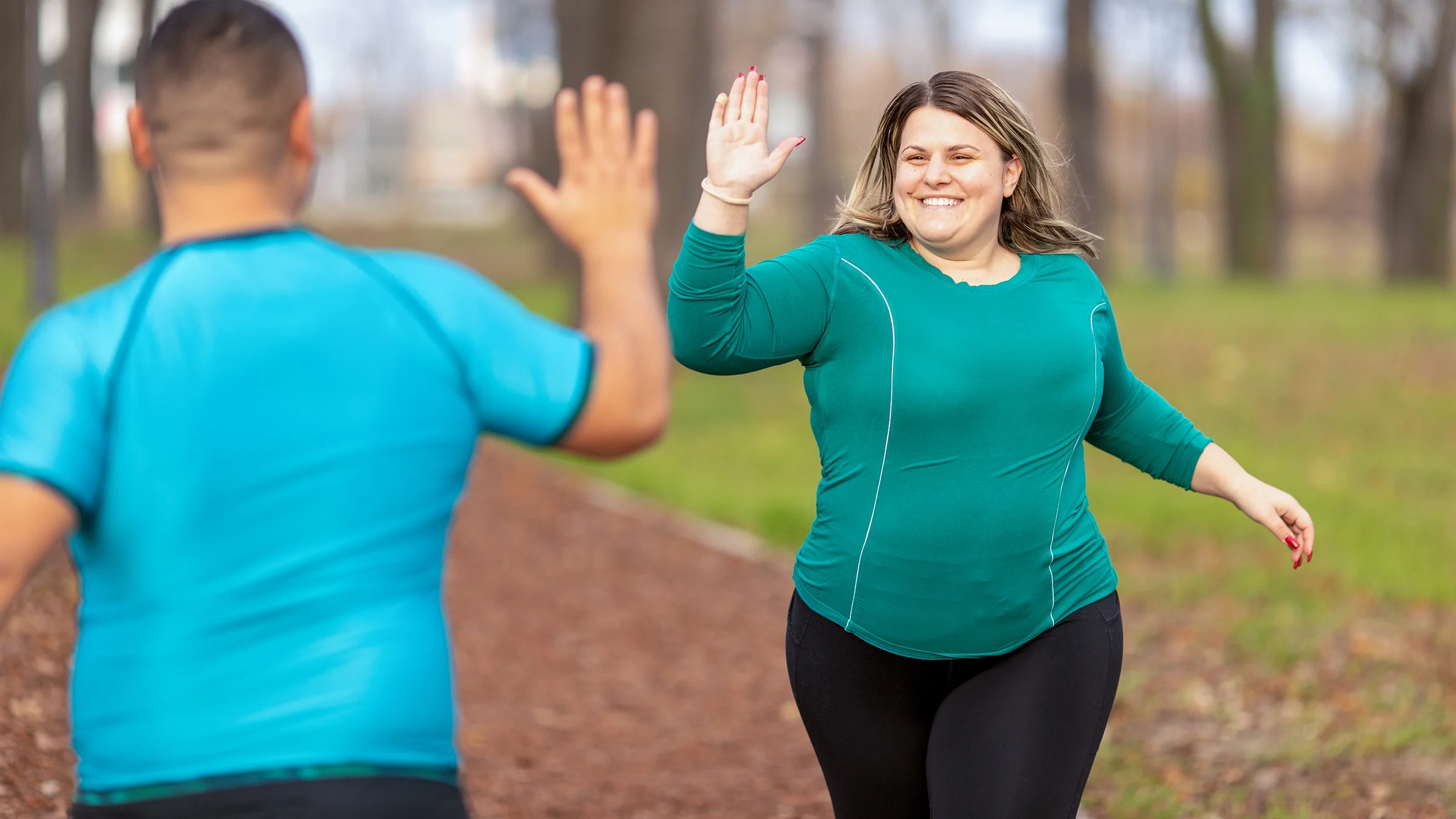 Two people on a run through the park passing each other. They are high five-ing as they pass.