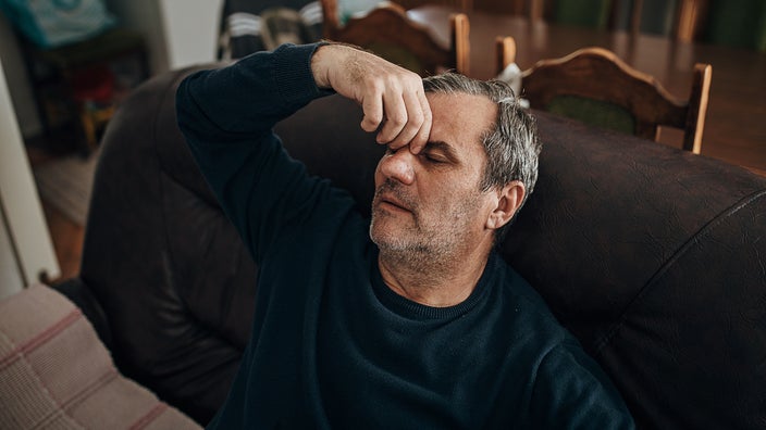 Man sitting on the couch at home pinching his nose and closing his eyes. He looks like he is in pain.