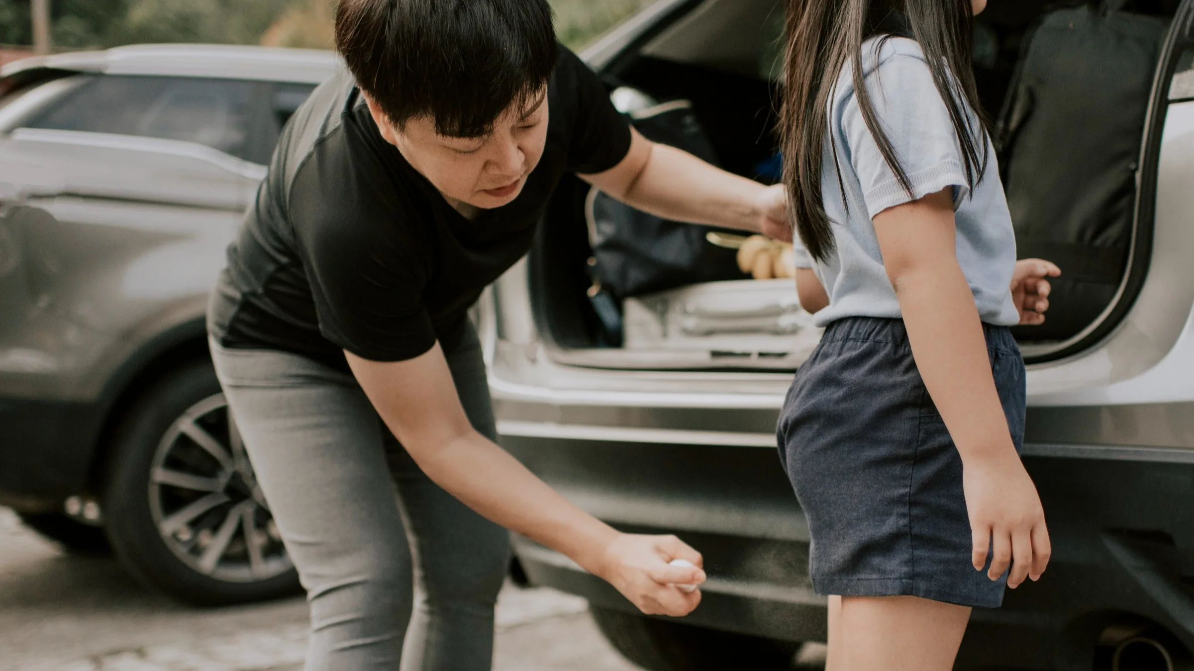 Mothering spraying the back of her daughters exposed legs with bug spray by the car before a hike.