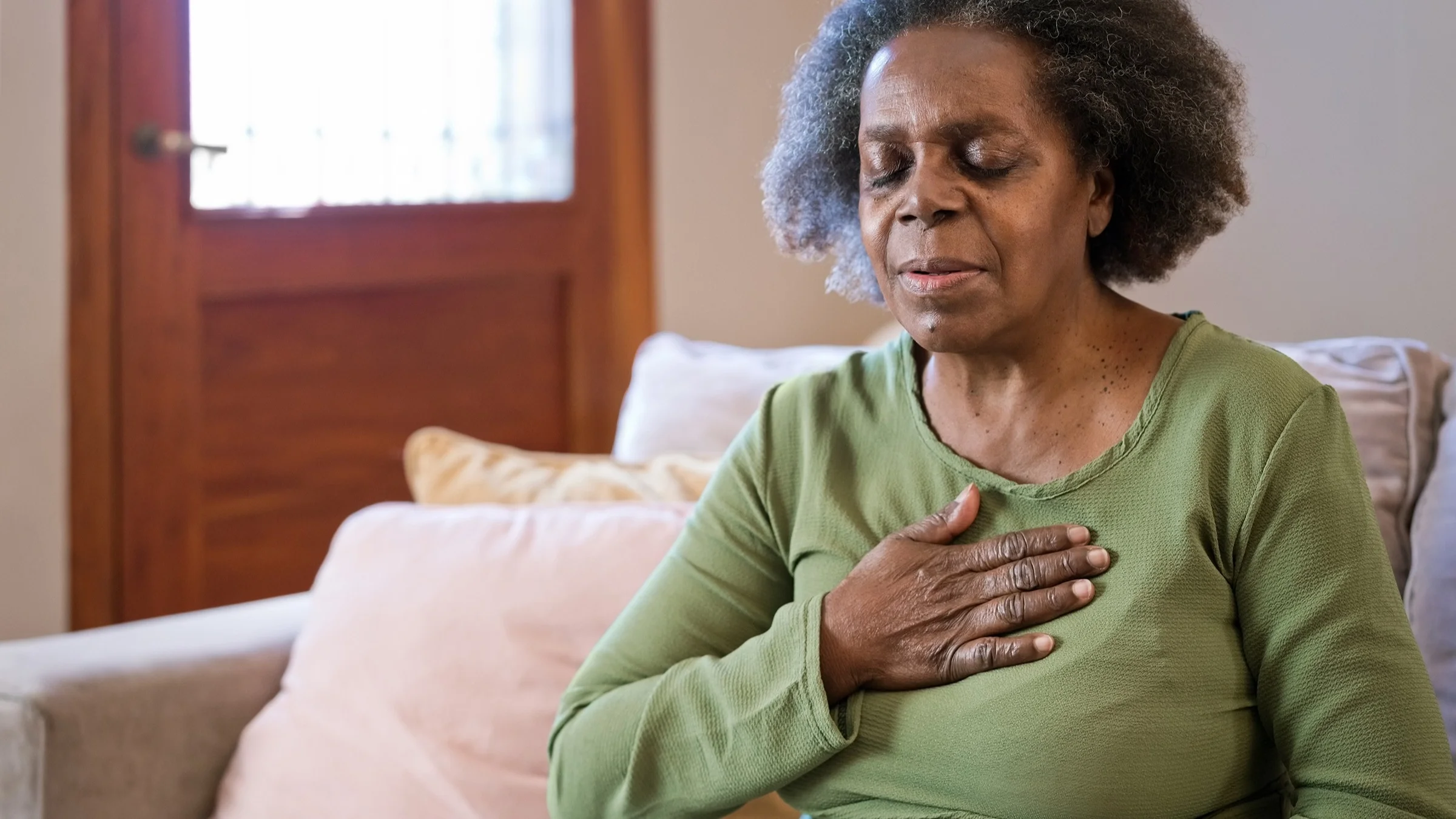 Elderly woman having chest pain. She has her hand resting on her heart as she has her eyes closed. She is wearing a green long sleeve shirt and sitting in her living room.