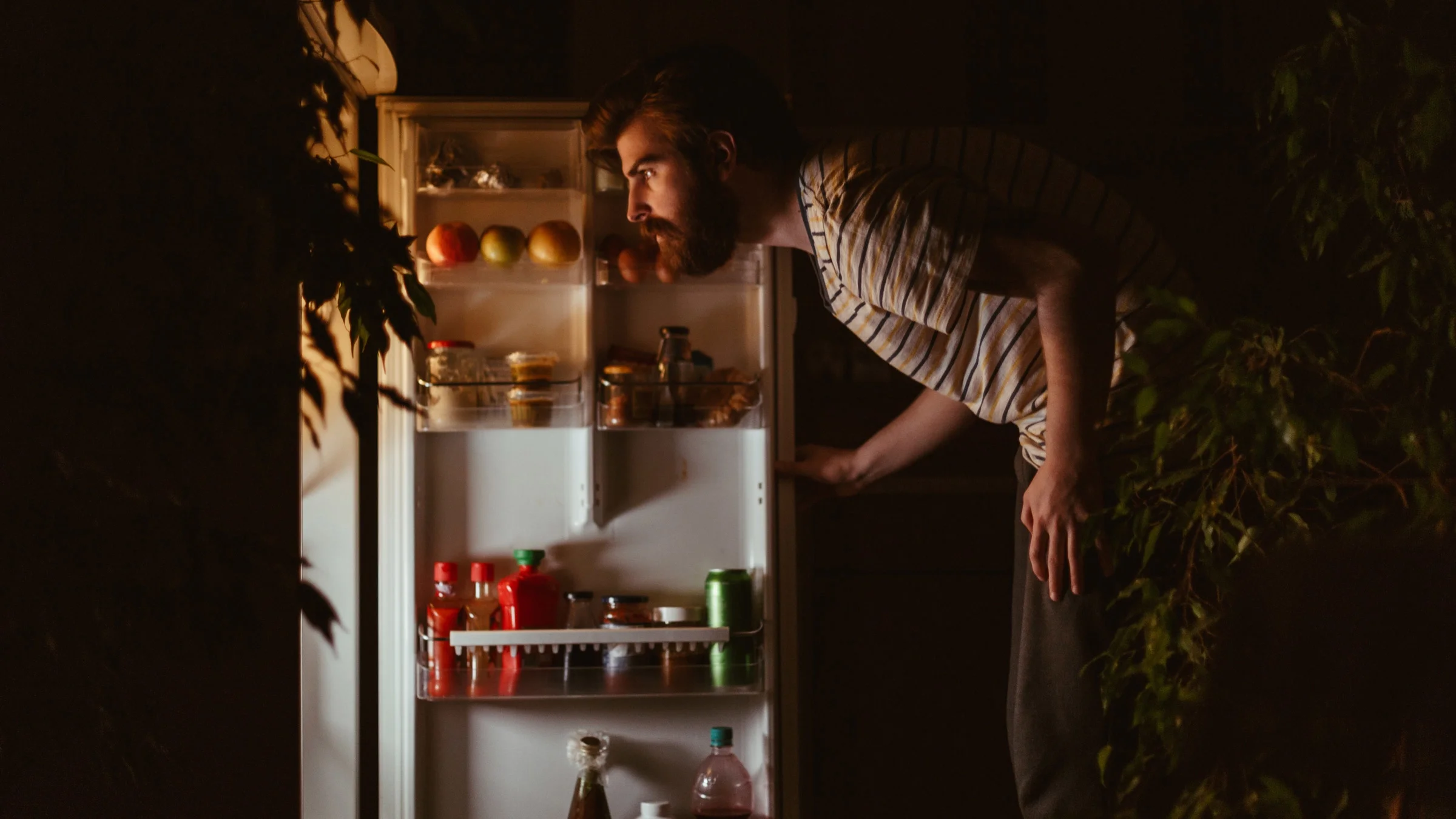 Red-head man looking into an open fridge in the dark. He is illuminated by the light coming from the fridge. 