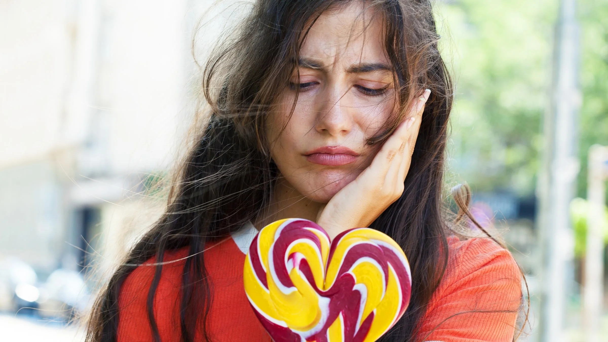 Holding cheek out of toothache, while holding heart-shaped lollipop. 