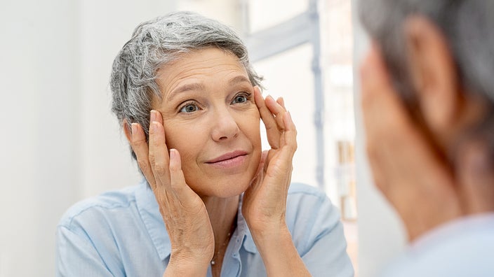 Older woman with gray hair looking in the mirror at her wrinkles on her face.