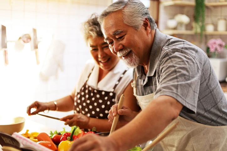 Pareja de adultos mayores cocinando juntos con delantales, sonriendo y riendo.
