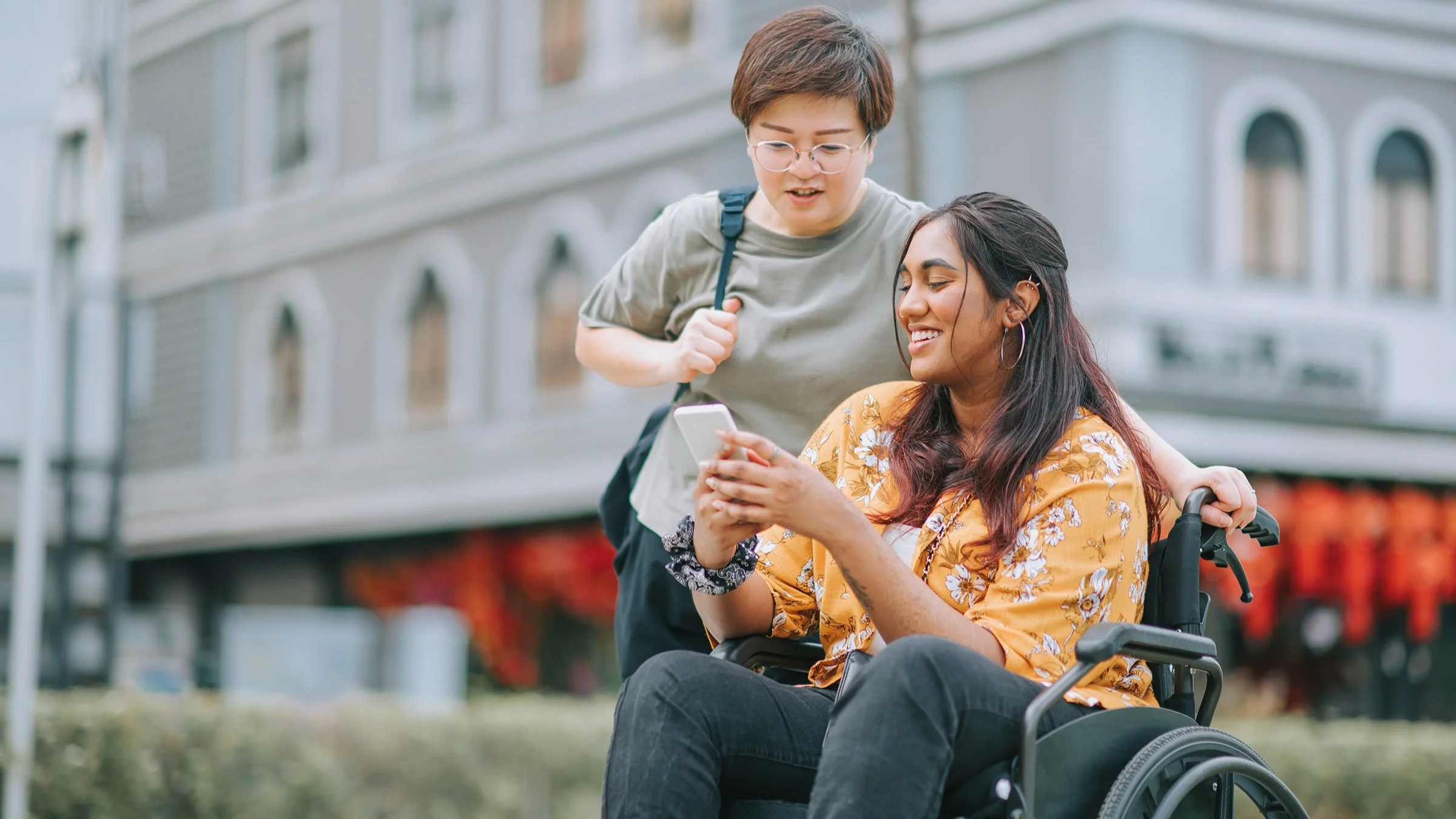 Friends traveling together through European streets. One woman is in a wheelchair. She is showing her friend something on her phone as the friend leans in to see.