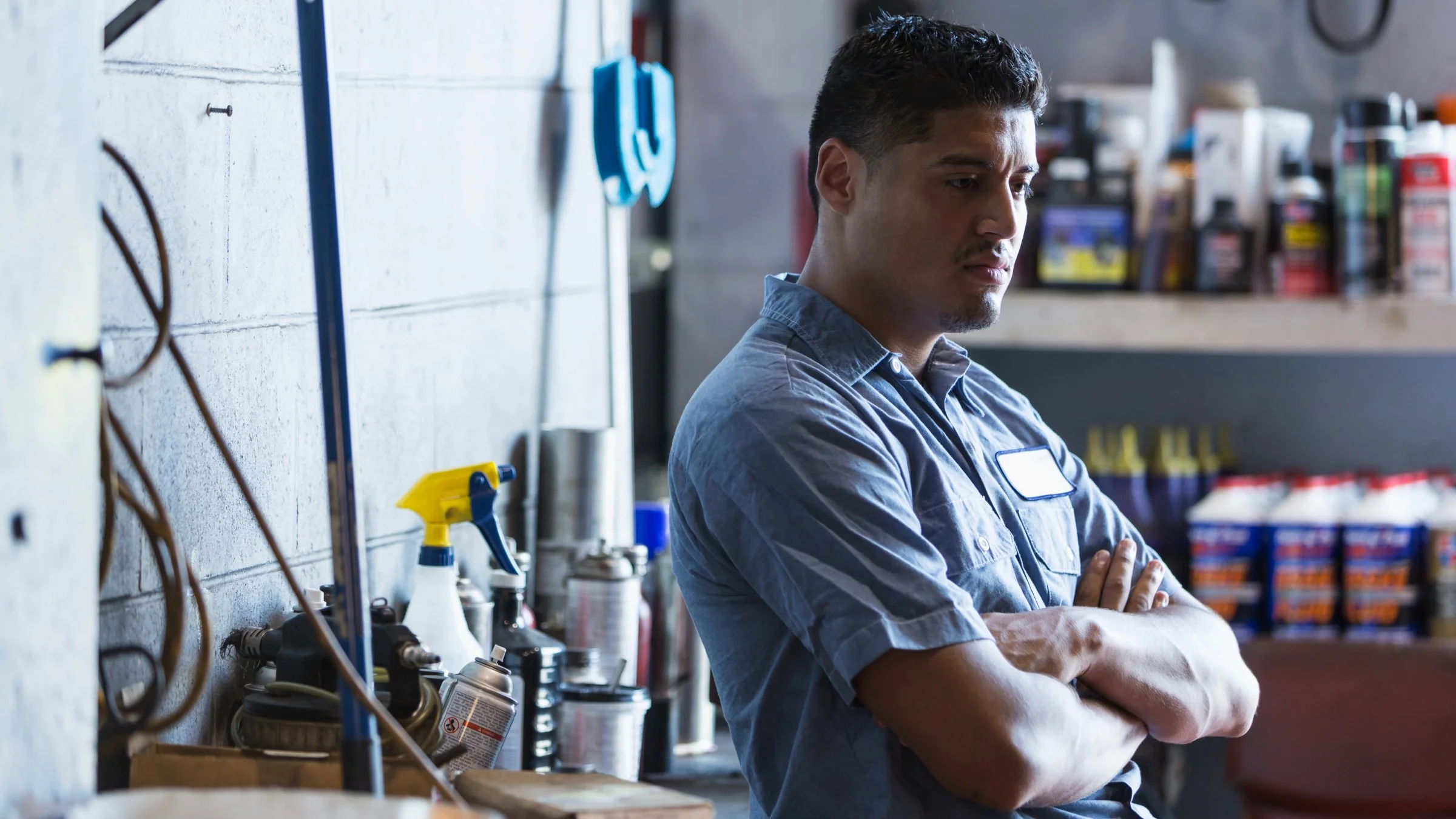 A mechanic worker leaning against a work bench looking down off frame with a sad despondent look on his face.