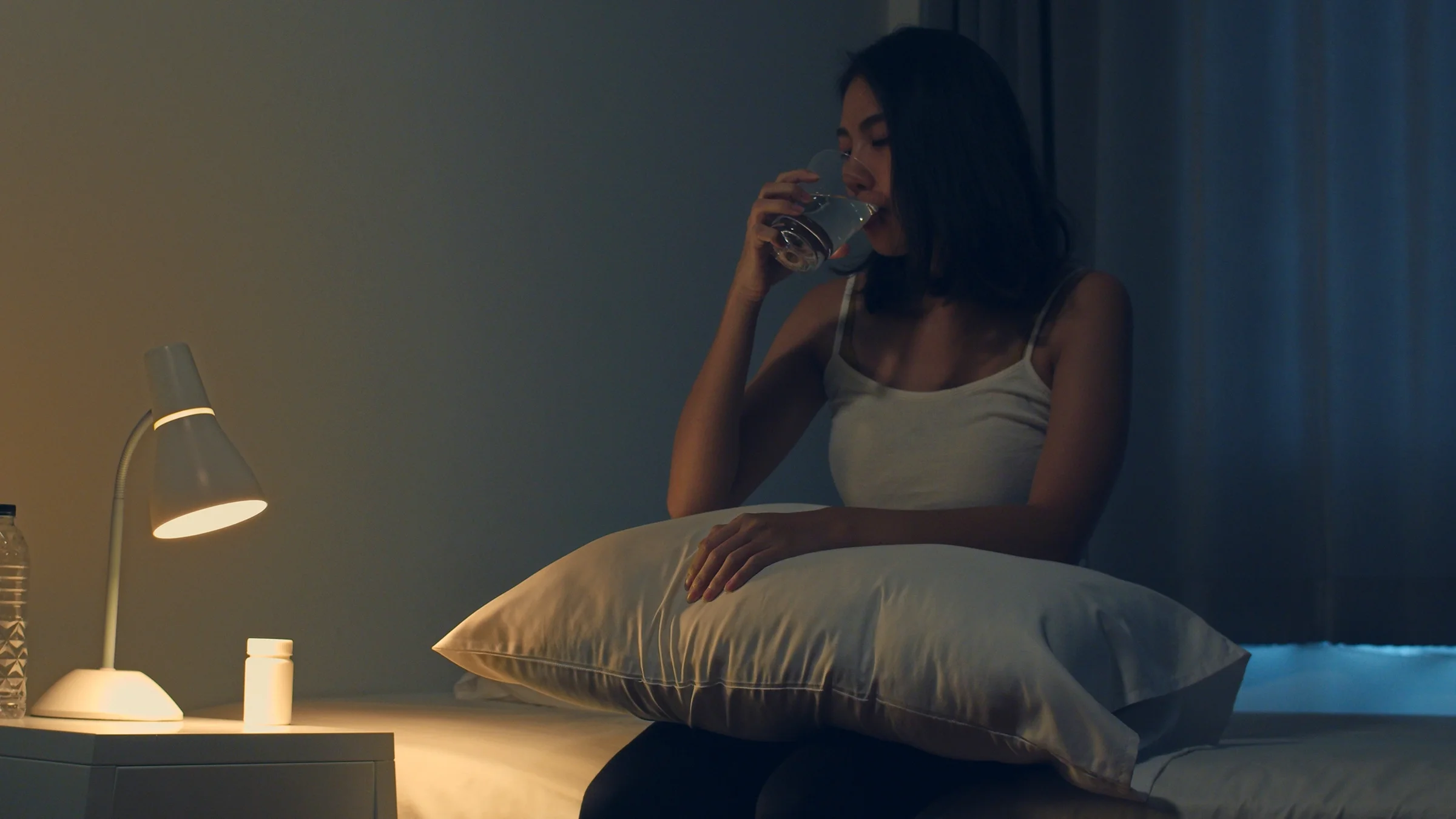 A woman sits on the edge of her bed as she takes a sleeping pill.