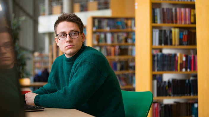 College aged man sitting in the school library zoning out looking away from his computer.