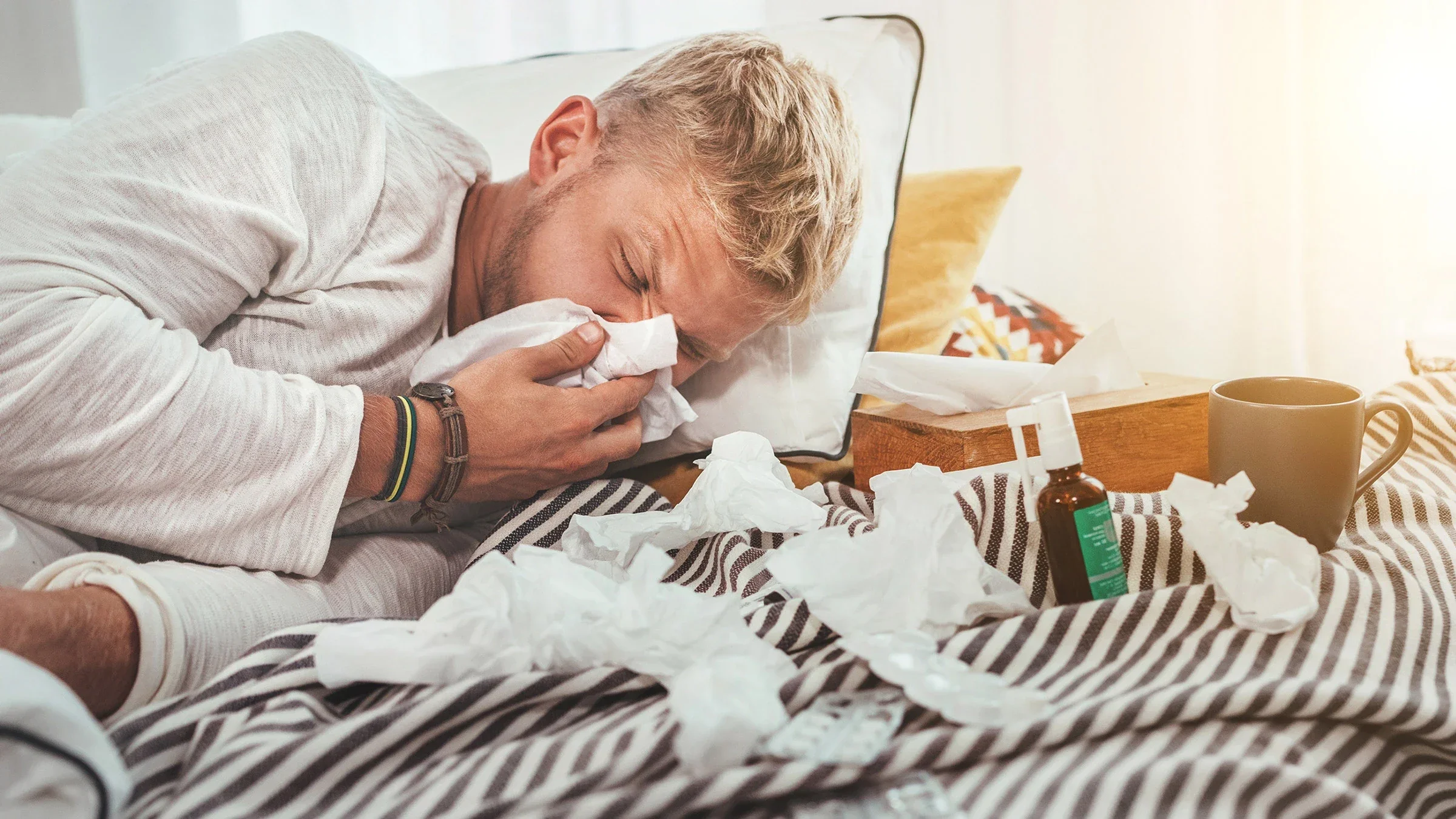 A sick person in bed blowing their nose and surrounded by used tissues and medicine 
Solovyova/iStock via Getty Images 
