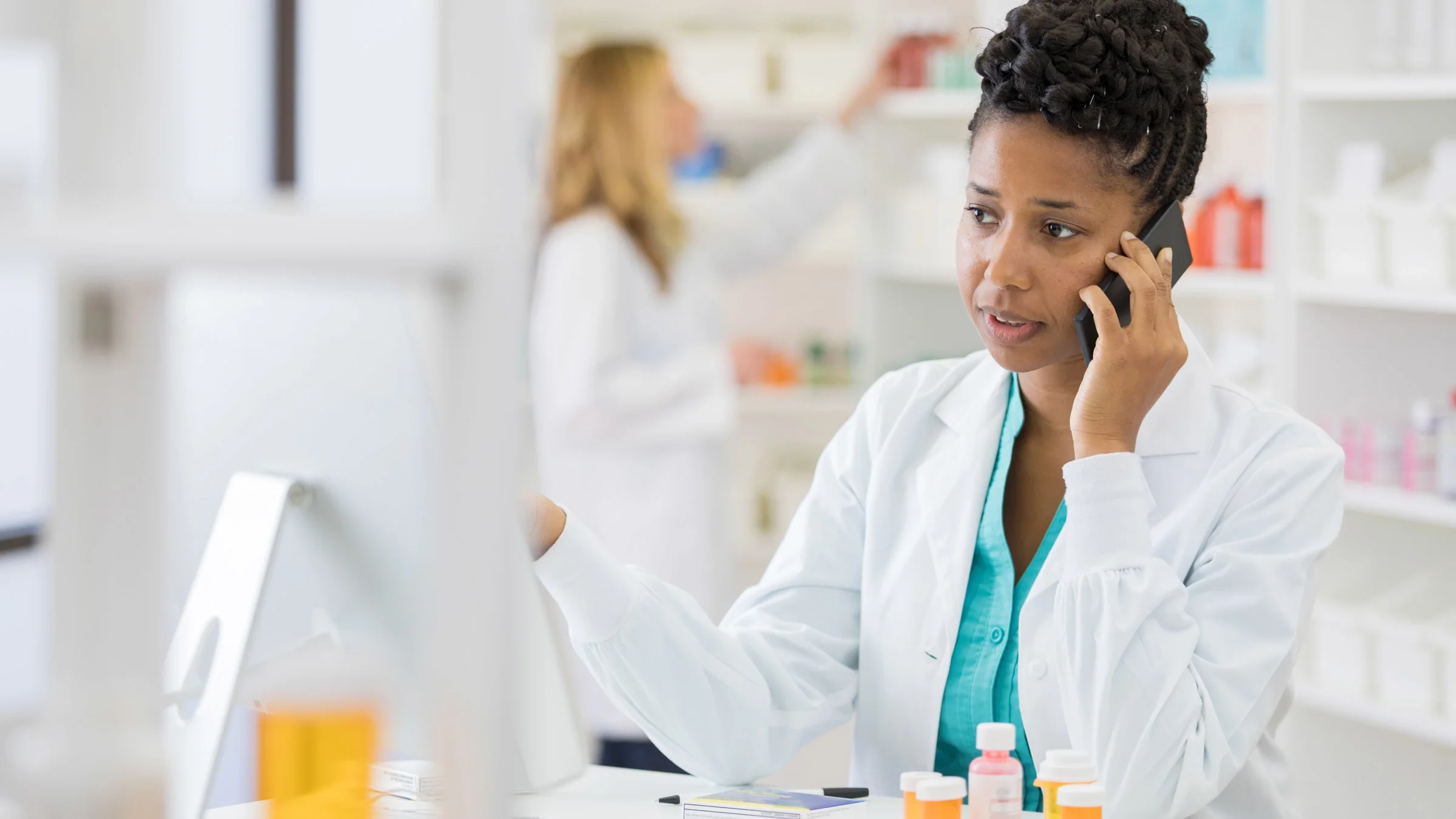 Pharmacist standing in front of her computer, talking on the phone. There are medications sitting on the table and the background is blurred.
