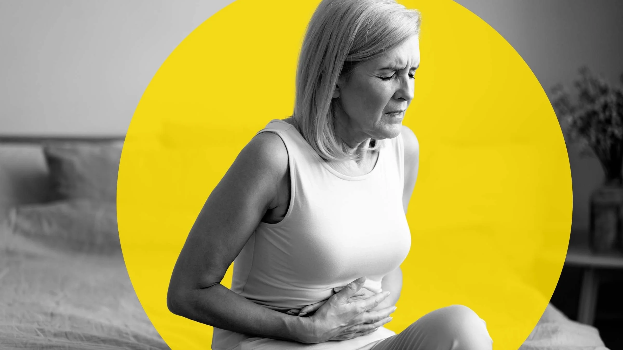 Black-and-white photo of a middle-aged woman with stomach pain. She is wincing and grabbing her stomach. There is an added yellow graphic circle added behind her as well.