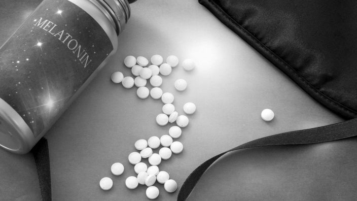 Black and white photo of a melatonin bottle with pills laid out on a plain background. There is also a silk eye mask in the top corner of the frame.