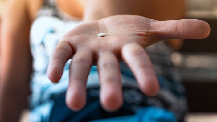 Close-up of a woman's hand holding out a small white pill to the camera.