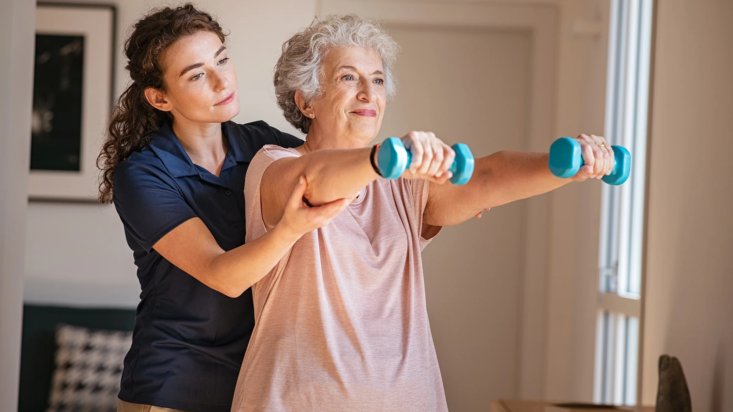 Senior woman lifting weights while doing physical therapy with a nurse at home.