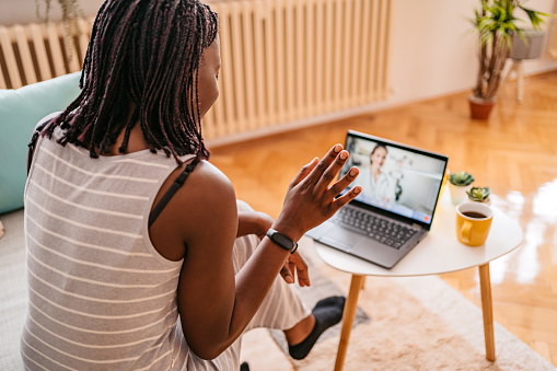 A young woman sitting on her couch waving hi to her doctor on a telehealth visit on her laptop.