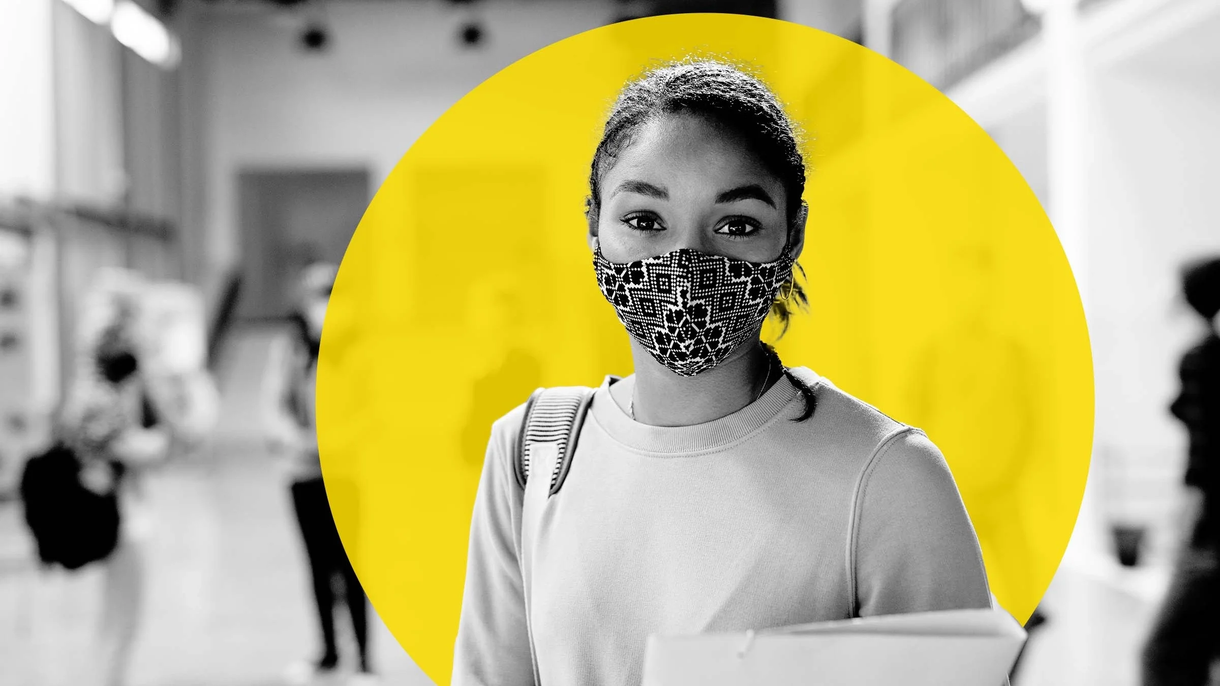 Portrait of a high school student standing with her binder and backpack. There are other students in the background social distancing. All are wearing face masks.