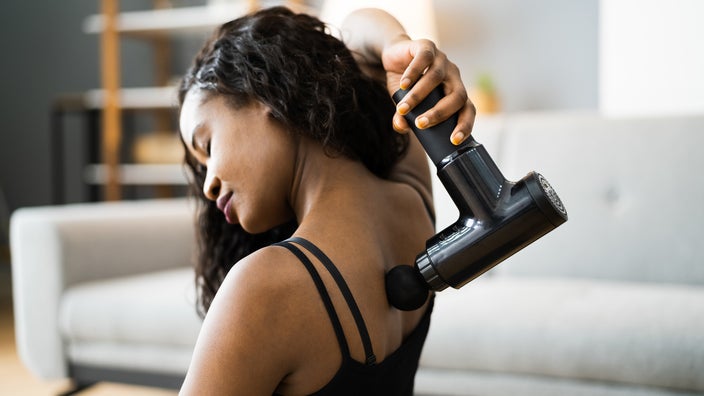 Young woman in workout clothes using a massager gun on her upper back.