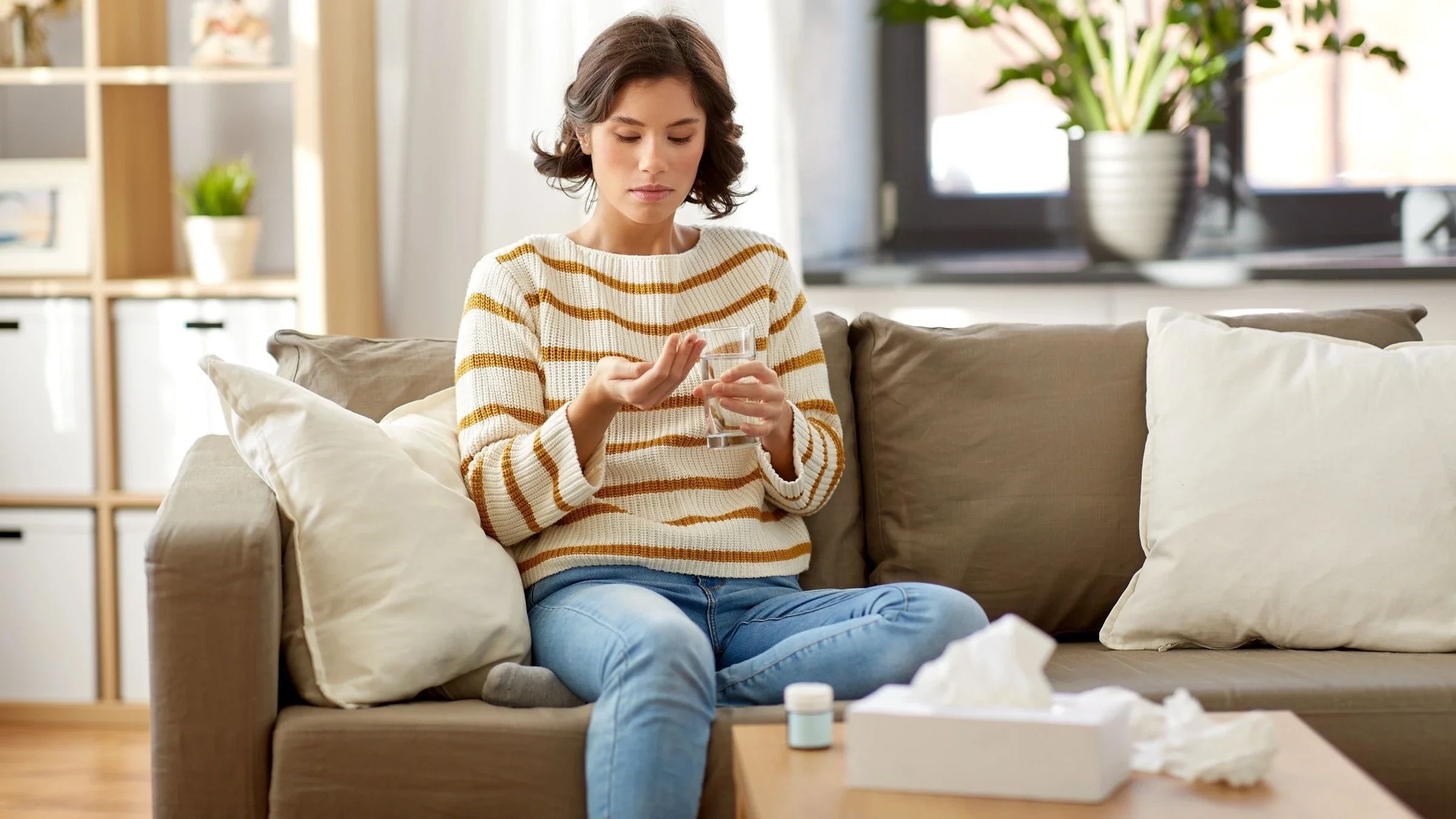 Young woman with short brown hair sitting on the couch taking medicine with a glass of water surrounded by used tissues.