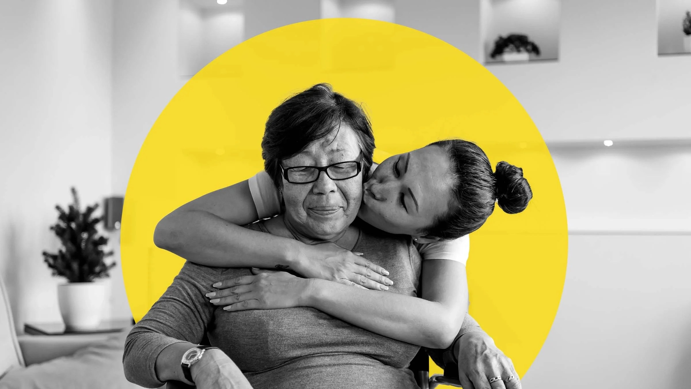 Black and white image of a young woman hugging her mother from behind as she sits in a wheelchair. The young woman is also kissing the mother on her cheek. There is a graphic yellow circle added behind them as well.