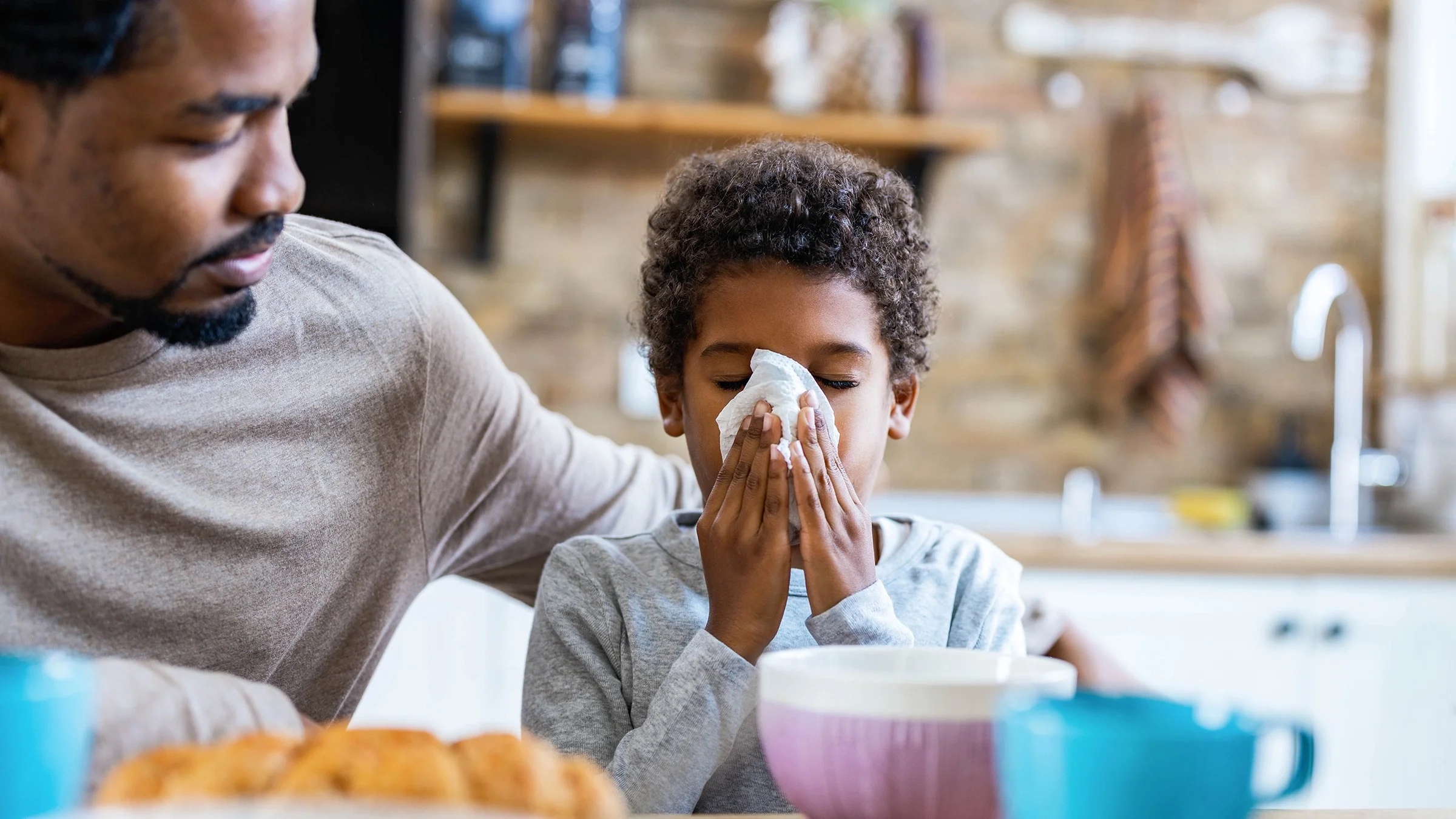 Portrait of a young boy blowing his nose with his father sitting next to him at the kitchen table. The father is comforting the young boy with his arm around his son's shoulders.