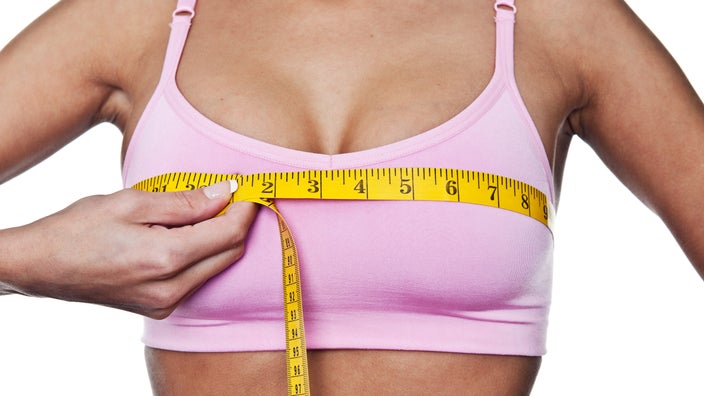 Close-up of a woman in a pink sports bra, she is using a yellow tape measure to measure her chest. 
