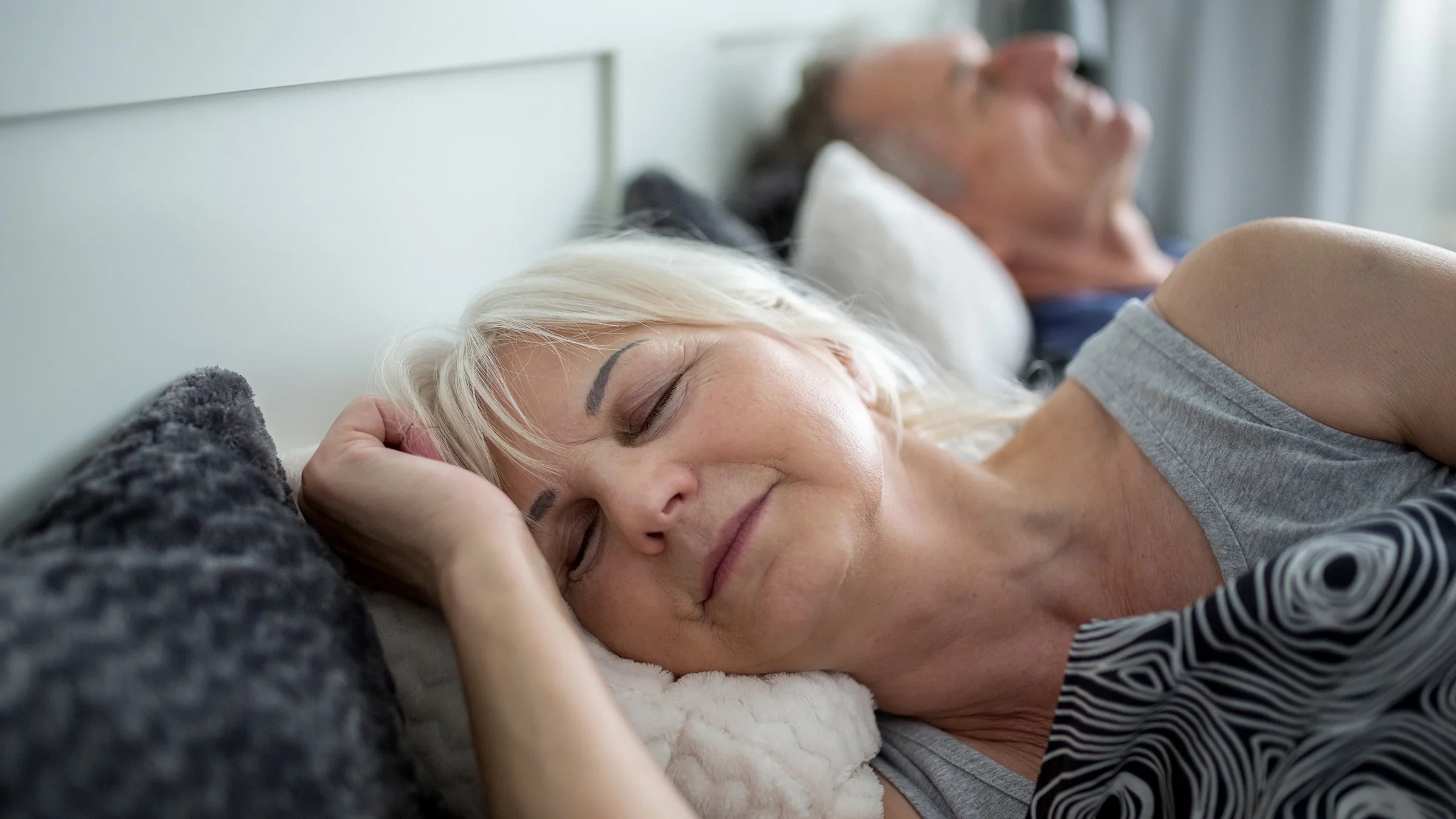 Close-up of a senior woman with white hair sleeping. You can see her husband on the other side of the bed in the background out of focus.
