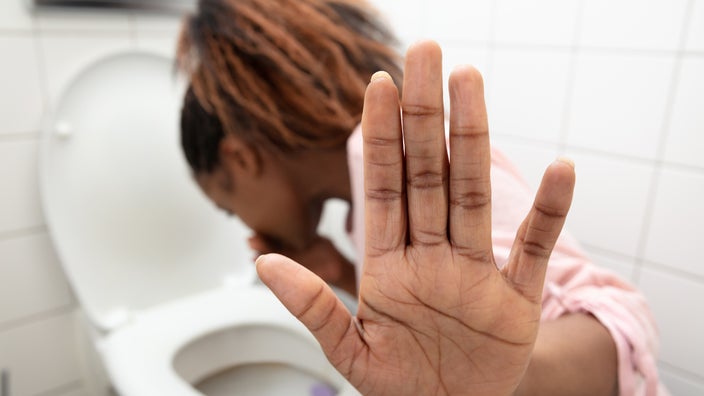 A woman is putting her hand up to a camera to stop the view of her vomiting into the toilet.