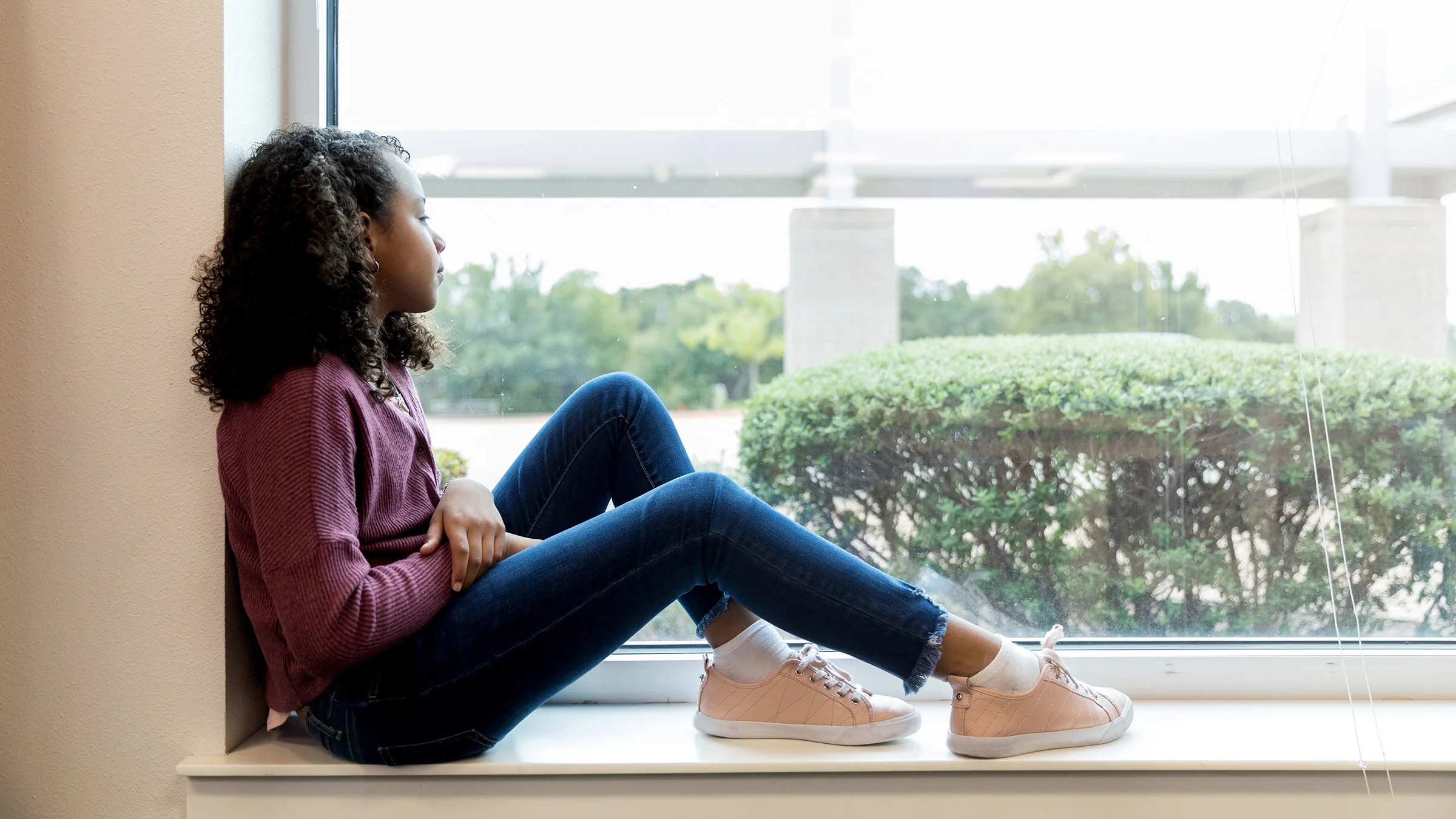 A young girl is sitting by a window looking out with a sad expression.