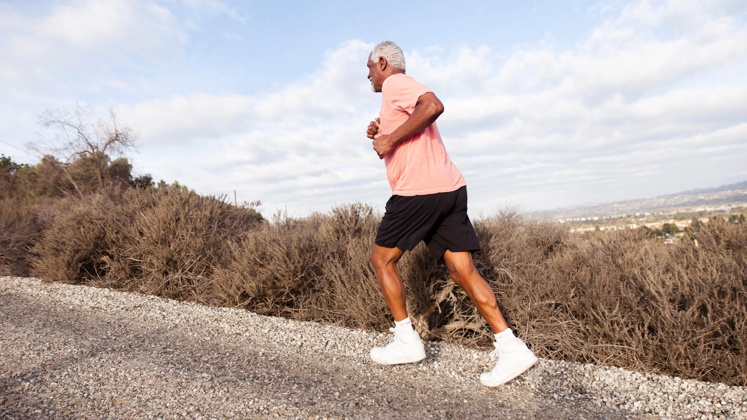 Elderly man jogging up a trail. He is wearing black running shorts and a pink t-shirt.