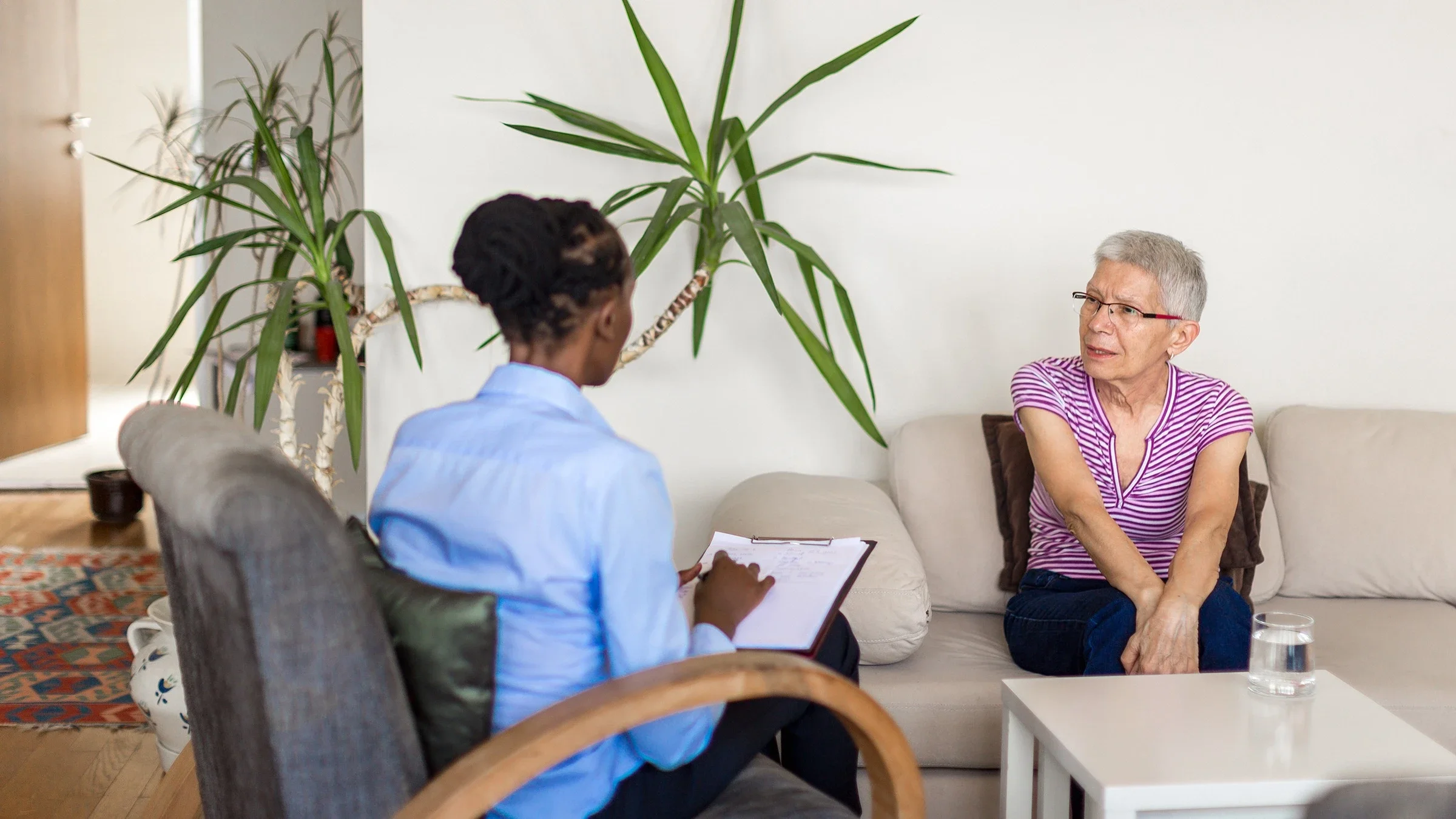 Senior woman at her therapist appointment. The therapist is sitting in a gray chair taking notes on a clipboard.