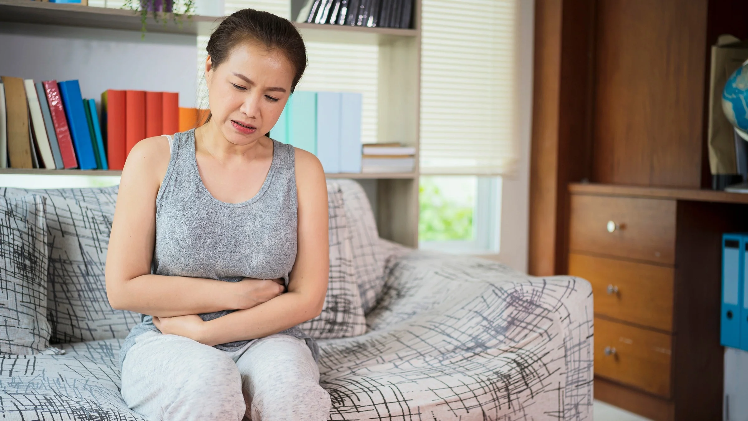 Woman sitting on the couch clutching her stomach in pain. She is wearing a gray tank top.