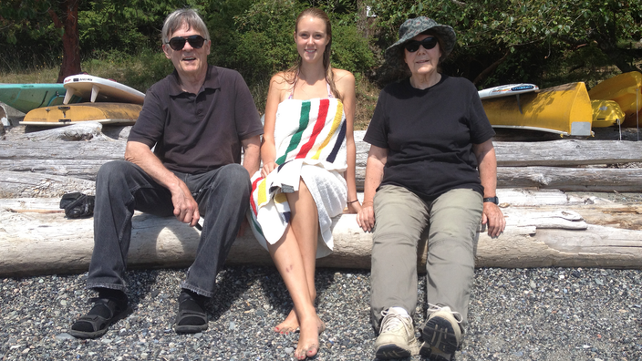 Author Clare Hennig, wearing a swimsuit and wrapped in a towel, sitting between her grandparents on a beach. 