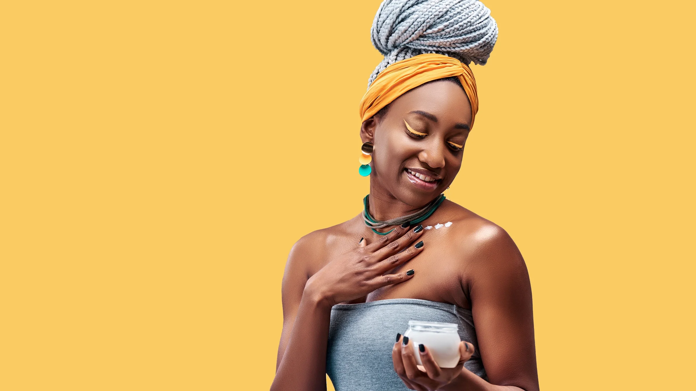 Portrait of a stunning beautiful woman with braids up in a top knot. The focus is her putting cream onto her neck and body. She is on a plain yellow studio background.