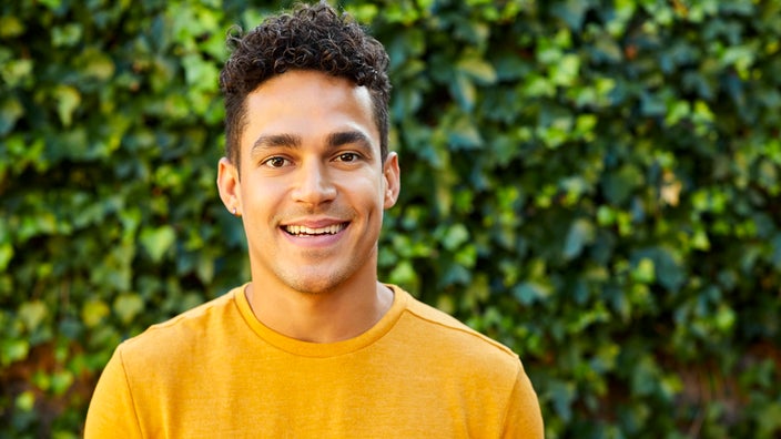 Portrait of a young man in a yellow shirt. The background is a lush green ivy wall.