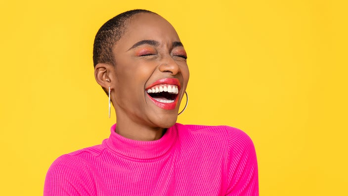 Portrait of a young woman who is extremely excited and laughing. She is wearing a bright pink mock turtleneck. The background is a plain yellow studio backdrop.