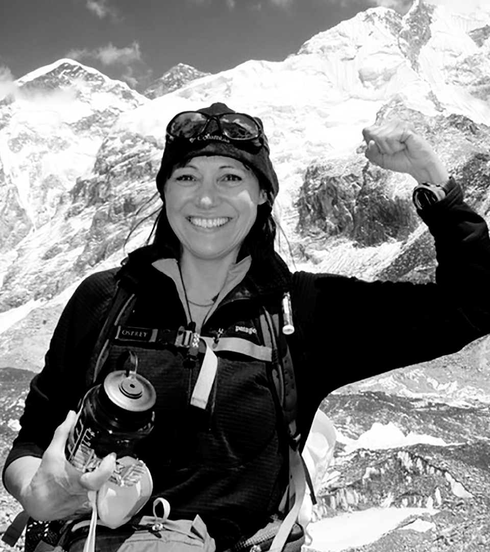 Black and white photo of Lori Schneider victoriously flexing her arms on top of a mountain.