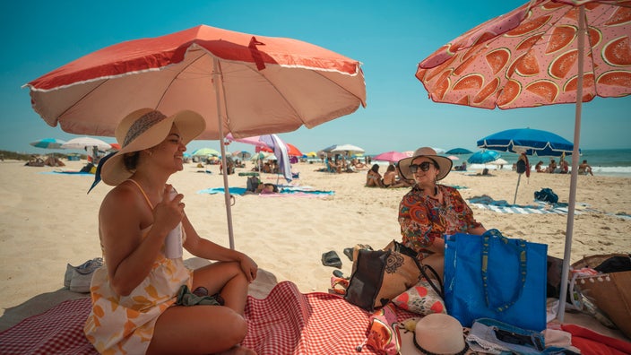 Two tourists wearing hats sitting under parasols on the beach.
