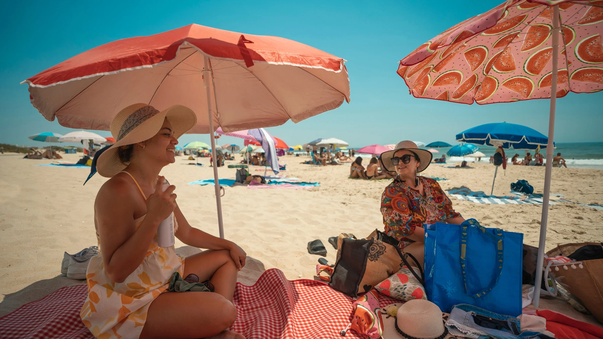 Two tourists wearing hats sitting under parasols on the beach.