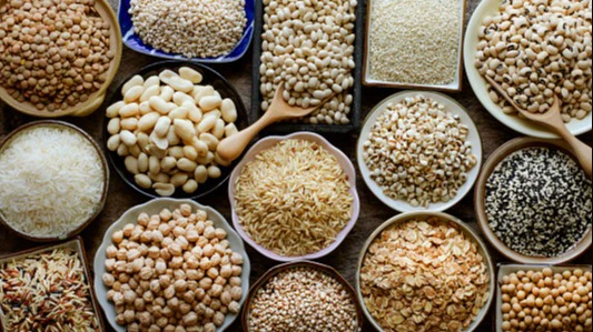 Top down view of a variety of different kinds of seeds, lentils, wheat, beans, and other grains in round containers on a wood table. 
